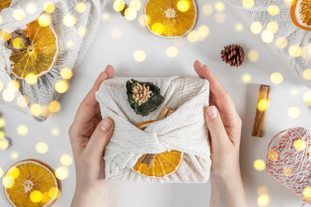 Gift Box in White Fabric. Female Hands wrapping Sustainable Gift on white Table with Decoration...