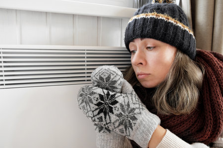 Young frozen woman wearing a sweater shaking and freezing for winter cold with shock expression on face. Girl is warming up hands with breath over electric heater. Discomfort spending time at home.の写真素材
