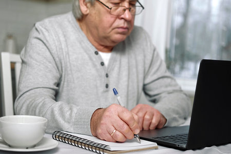 Senior man E-learning writing in notepad. Old male studying at home sitting at desk using laptop. Closeup hand writes in notebook with pen make note information. Older people distance learning conceptの写真素材