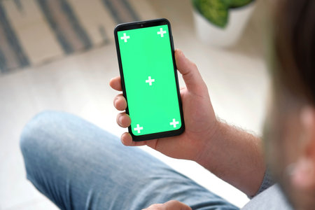 Young man sitting at home holding smartphone green mock-up screen in hand. Male person using chroma...