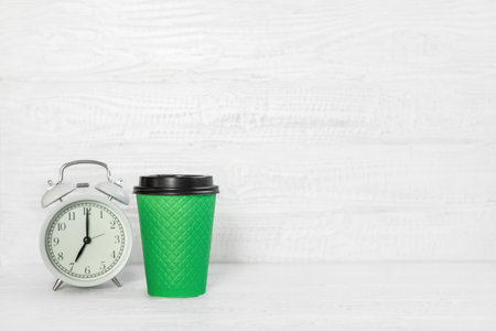 Front view white desk with alarm clock, coffee paper cup, copy space. Morning background banner with free space for text. Minimalistic concept, workspace, time to work. Blank workplace. office mock-upの写真素材