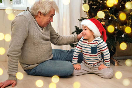 Grandpa and Little Boy at Christmas Time. Grandfather and Grandchild on the Floor near Christmas Tree. Winter Holiday Concept. Hoary Old Man Hiding a Gift for Small Toddler Child. Celebrating Togetherの写真素材