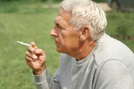 Senior man lighting up a cigarette outdoors against green nature background. Old thoughtful man smoking, looking aside. close up face. Relaxed pensioner enjoy weekend rest outside. unhealthy habitsの写真素材