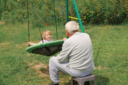 Grandfather swinging a child in summer at sunset. Grand dad and grandchild sitting on swing in the park. Senior 60s Grandpa pushing beautiful baby on the rope seesaw. old man and little girl at playgroundの写真素材