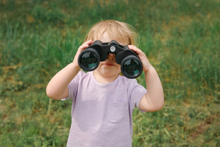 Little girl using binoculars in a forest. 2 years kid looking ahead. Smiling baby with spyglass. Adventure, Imagination, travel concept. Freedom vacation. Happy child playing outdoor in summer fieldの写真素材