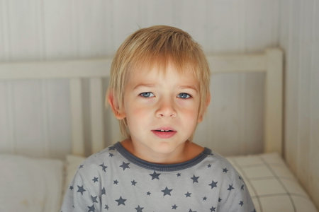 Portrait of 5 years Boy in pajamas in the light bedroom. Child making face. Funny foolishness portrait of little boy in white bed. Adorable caucasian kid looking at camera doesn't want to sleepの写真素材