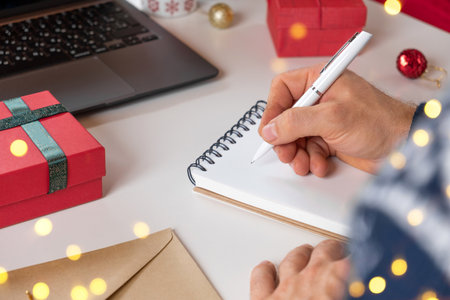 Writing a Christmas Wishing Card. Male adult making greeting cards at home office during Xmas. Man hands in blue sweater holding blank paper and envelope at the Desktop. Coffee Cup, holiday decorationの写真素材