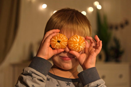 Merry Christmas. Funny child boy laughing and dancing covering his eyes with tangerine. Happy New Year. Portrait of excited little kid wearing pajamas having fun at home on Xmas night. Festive moodの写真素材