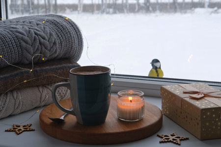Winter windowsill still life. Blue ceramic cup of hot coffee on window sill. Christmas decorations on the background. Cozy home picture. Warm woolen knitted sweaters, Burn Candle, Cookies. Stock photoの写真素材