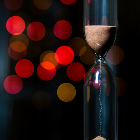Hourglass in the center. Sand strews down on christmas background, bokeh red christmas lights. Time...