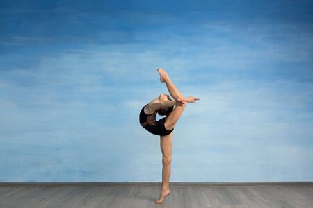 A young girl in a black gymnastic swimsuit gymnast makes an exercise standing backward and legs in semi splits. A young girl gymnast performs kick back arms straightened backの写真素材