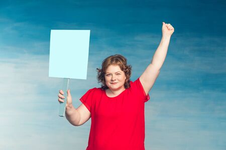 Cute smiling chubby red-haired woman screaming fervently, waving his hand in the second holds a protest poster on a blueの写真素材