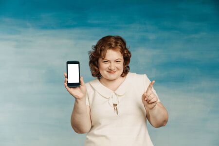 Cool red-haired plump woman in a white dress exults with a phone in her hands on a blue