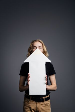 A young guy with long blonde hair is holding in front of his face a white arrow pointing upward on a...