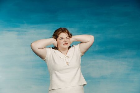 A plump young woman in a white dress with red hair covered her ears with her hands from annoying...