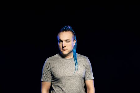 A man with a braided mohawk haircut calmly posing holding hands in pockets of sweatpants against a...