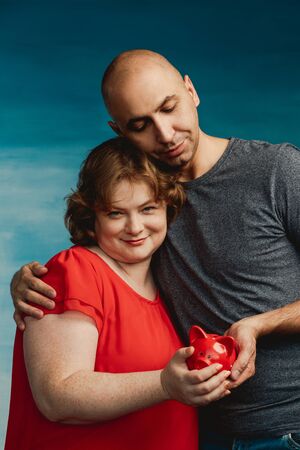 Man and woman holding a piggy bank and puts a coin in a red piggy bank which is on the male hand against a blue background.の写真素材