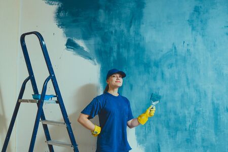 A young woman in a blue T-shirt in yellow rubber gloves and cap is painting a wall in an apartment,の写真素材