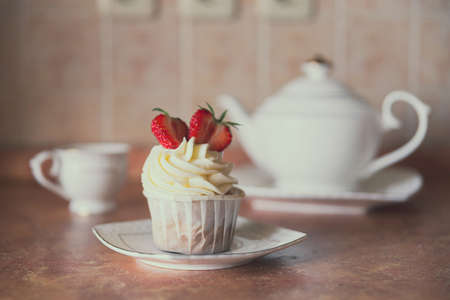 Appetizing cupcake with white cream decorated with fresh strawberries on the table, accompanied by a white teapot and cup.の写真素材