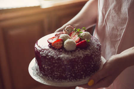 Womens hands are holding a mousse cake covered with pink icing, decorated with a figurine of a...