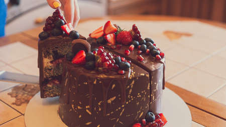 The hand cuts off a piece of appetizing chocolate cake. chocolate decorated with strawberries, blueberries and a pomegranate on an orange backgroundの写真素材