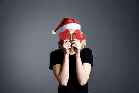 Young guy an attractive guy with long blonde hair in the form of a Christmas elf in a Santa Claus hat closed his eyes with red hearts on a gray background.の写真素材