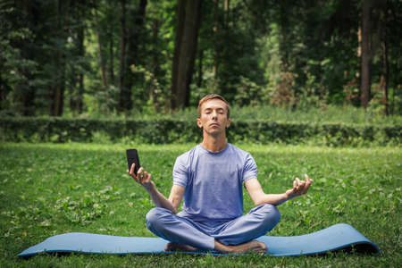 Young attractive white man learns to meditate in the park in front of smartphone.の写真素材