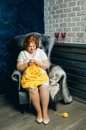 A beautiful plump redhead young woman with freckles sits in an armchair and knits a yellow sweater.