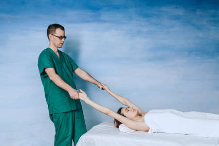A male massage therapist massages a young woman, a method of stretching arms on the blue background