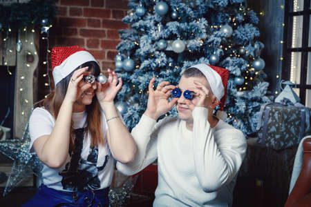 A young man and woman in Santa Claus hats are fooling around on the background of the New Year tree