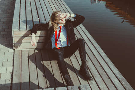 Portrait of a young man in a suit sitting on a wooden bridge and covering his eyes with his hand...