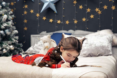 A girl in pajamas lies on the bed and writes a letter to Santa Claus or grandfather frost on the background of a Christmas treeの写真素材