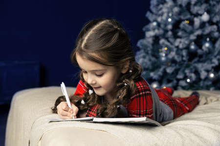 A girl in pajamas lies on the bed and writes a letter to Santa Claus or grandfather frost on the...