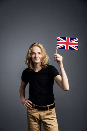 A young guy with long blonde hair in a nuclear t-shirt smiles and waves the UK flag on a gray background.の写真素材