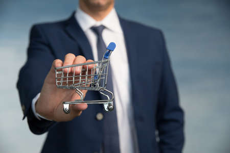 Male hand of a businessman in a blue suit with a tie hold a mini shopping trolleyの写真素材