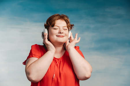 Young attractive plump woman with red hair listening to music with pleasure headphones with eyes...
