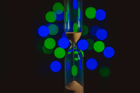 Hourglass in the right. Sand strews down on christmas background, bokeh blue, green, violet lights. Time flow. Time is overの写真素材