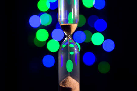 Hourglass in the right. Sand strews down on christmas background, bokeh blue, green, violet lights....