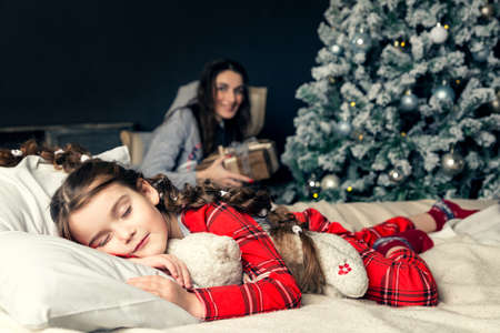 The girl sleeps on New Year's Eve, hugging a soft toy, while mom puts gifts under the Christmas...