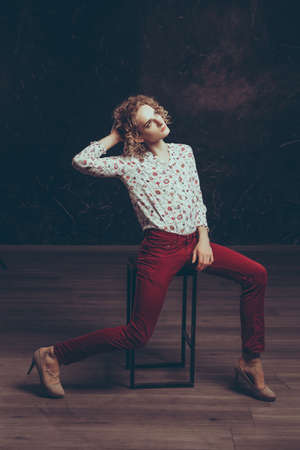 A young blond handsome man androgyn in the image of an attractive woman posing sitting on a chair on a dark backgroundの写真素材