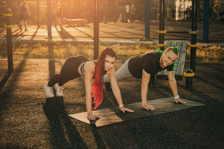 A man and a woman with interesting hairstyles do a plank exercise on a city sports ground.の写真素材