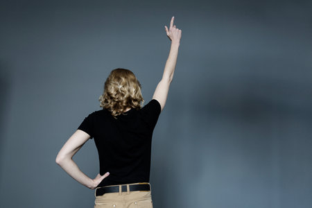 A thin young man with long blonde hair in a black T-shirt and beige jeans, stands with his back to the camera and shows thumbs upの写真素材