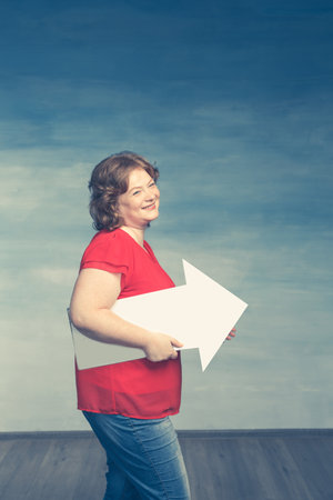 Pretty red-haired plump woman in a red T-shirt and jeans holds a white up arrow to the side on a blue backgroundの写真素材