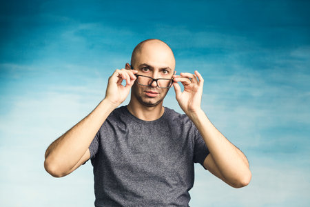 An interesting bald man adjusts his glasses with his hands and looks in surprise at the camera on a blue background.の写真素材