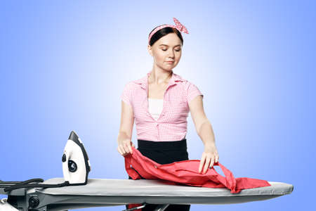A beautiful girl dressed in pin-up style is ironing clothes isolated on a blue background.の写真素材