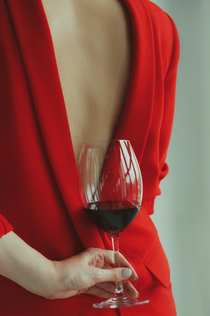 A young woman in a red jacket holds a glass of red wine behind her backの写真素材