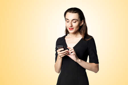 Business woman (fatal woman) in a black dress with red lipstick looks at her phone, isolated on a yellow background.の写真素材