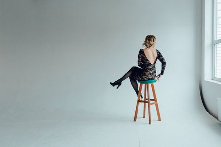 A young woman in a beautiful black guipure dress with an open back sits on a bar stool with her back to the viewer in an empty gray room.の写真素材