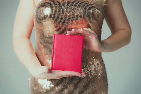 A girl in a gold evening dress holds a closed book with a red cover with two hands.の写真素材