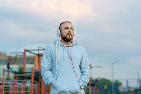 A man with a braid hairstyle in the early morning listens to music with headphones on the sports field against the background of the pink-blue sky.の写真素材